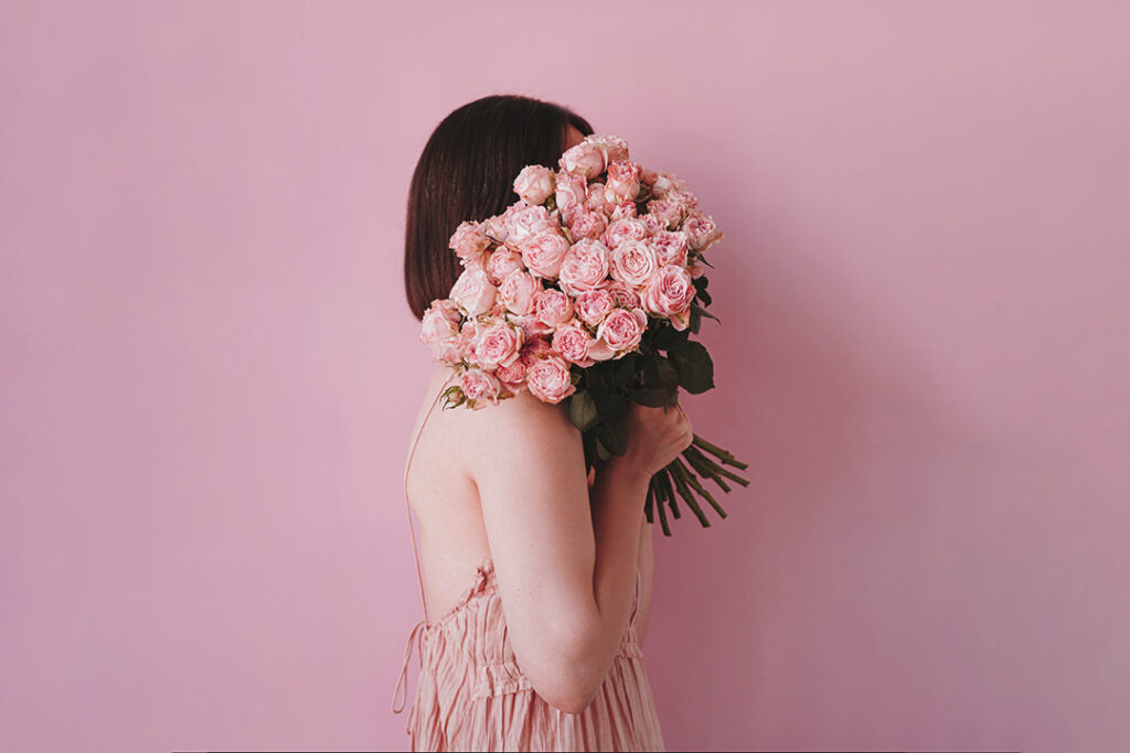 Eine junge Frau in der Seiten-Ansicht in einem rosa Kleid vor einem rosa Hintergrund versteckt ihr Gesicht hinter einem rosa Blumenstrauß - als Symbolbild für die Sichtbarkeit von selbstständigen Frauen - sie sind vom Typ her eher leise, bekommen aber überall gesagt, sie müssten laut sein, um "gesehen zu werden"